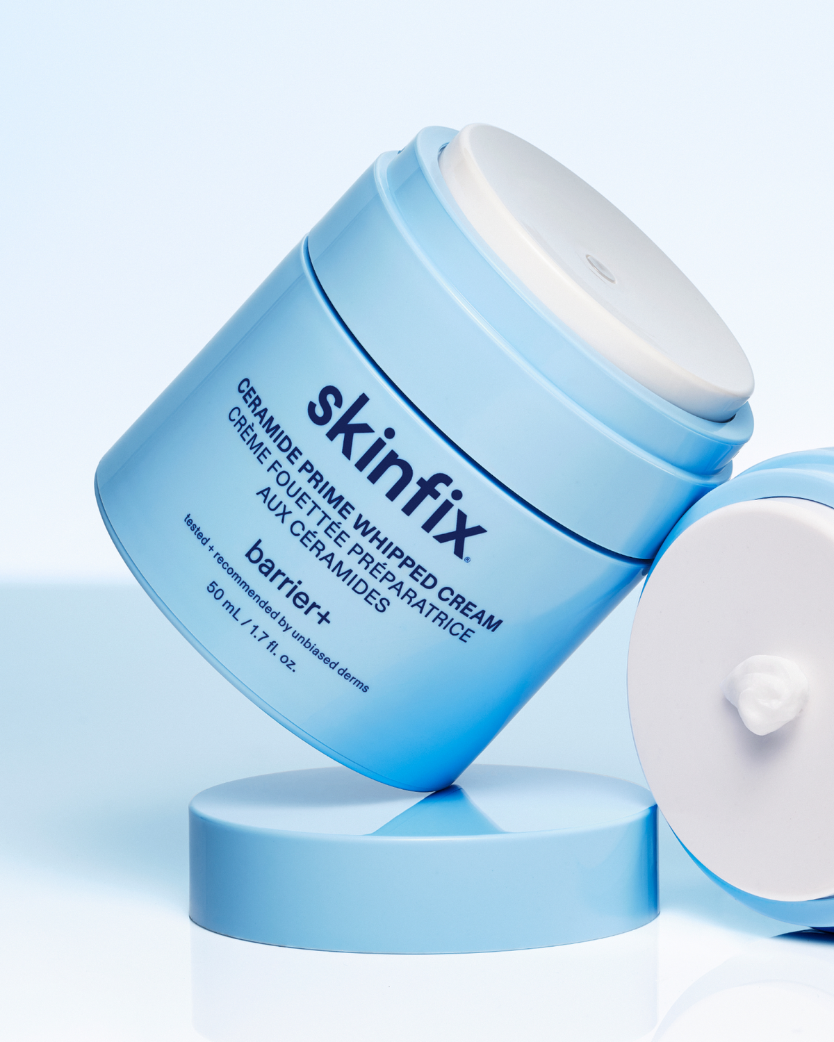 Skinfix Ceramide Prime Whipped Cream jar tilted at an angle on a blue pedestal, with the lid resting open beside it, against a light blue background.